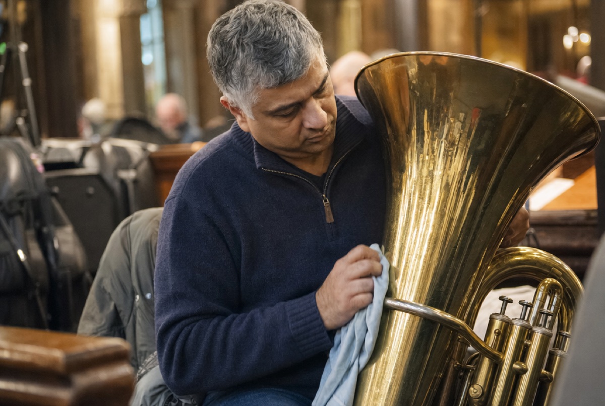 A band member polishes a large brass tuba on a chair in the church hall, instrument cases and music stands behind him.
