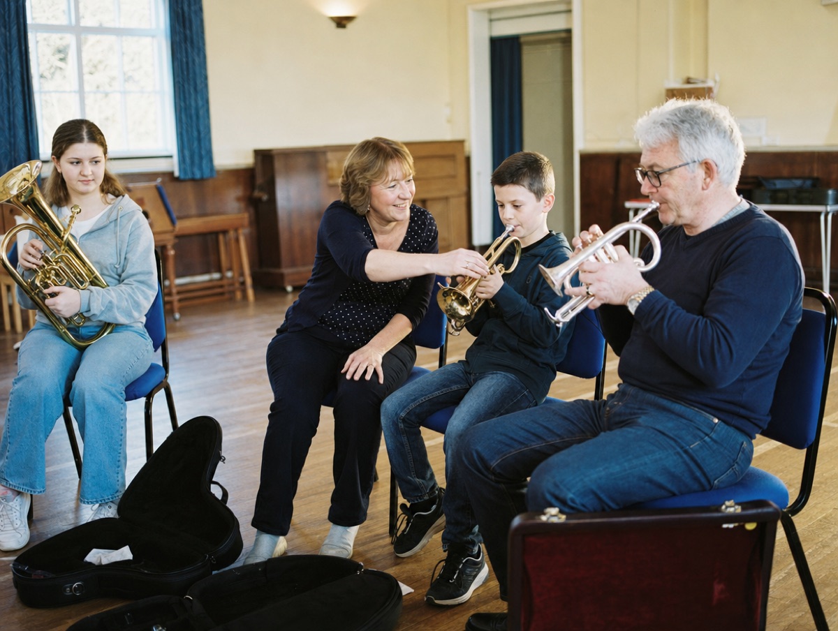 A tutor helps beginners of different ages hold their cornets at a training band session in a church hall.